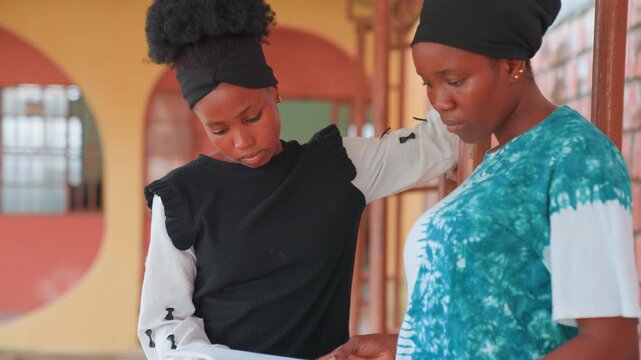 African mother and child reviewing paper in corridor, mother offers calm guidance while daughter reads document, warm brick background, quiet supportive moment, mother as counselor and daughter