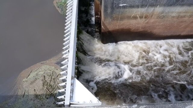 a horizontal high-angle video clip capturing turbulent white water rushing through an open sluice gate past a metal trash screen at a water control facility.