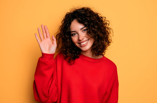 Attractive curly woman waved her palm to say hi or hello. Smiling broadly, she was greeting. Happy and friendly, the girl posed joyfully over the yellow background. Confident and feminine, the lady in