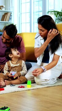 Indian Asian twin infant babies playing with colorful toys while young father and mother sit together on carpet in modern living room at home, capturing joyful family bonding and parenthood moments