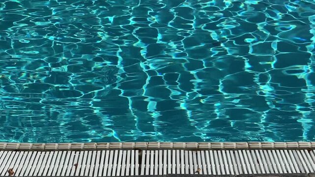 Blue swimming pool water surface with sunlight reflections and white plastic drainage grate at the edge. 4K.