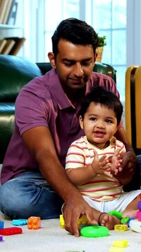 Indian Asian young parents playing with twin infant babies using colorful ring stacking toy at home, guiding fine motor learning activity and clapping with joy after completing playful task together