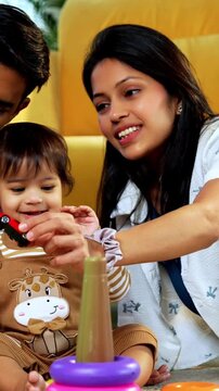 Indian Asian twin infant babies smiling and looking at camera while sitting with young parents on carpet surrounded by toys in modern home living room, capturing joyful family portrait and bonding