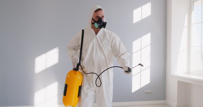 Exterminator spraying floor to kill insects, spray toxic chemical insecticide in empty room. Pest control worker with mask and suit using sprayer to apply pesticide for disinfection and fumigation