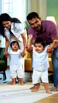 Indian Asian young parents helping twin infant babies walk by holding their hands inside modern home living room, guiding first steps together as smiling babies enjoy playful family bonding moment