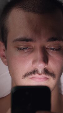 Top-down view portrait of a tired young Caucasian man lying in bed at night using his smartphone in the dark, suffering from social media addiction and insomnia.
