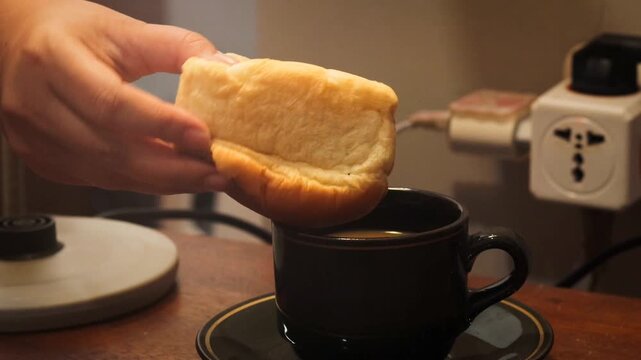 Asian woman's hand dipping a piece of bread into a cup of coffee at home. Simple breakfast and everyday dining activity.