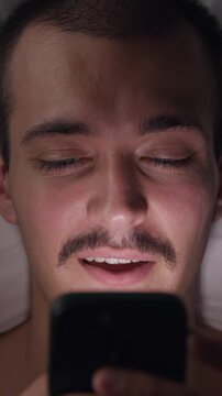 Top-down view portrait of a happy young Caucasian man lying in bed at night smiling and laughing while using his smartphone in the dark before sleep.