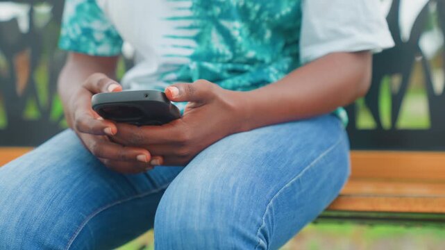 Black mother on bench checking phone, closeup of hands and smartphone, denim jeans and patterned top, natural light and soft bokeh, waiting to pick up child, texting and reading notifications