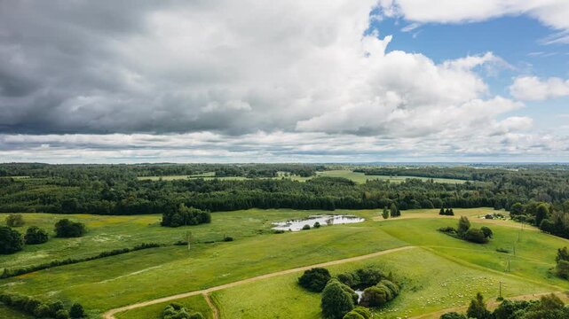 Aerial drone hyperlapse view of moving clouds over green rolling hills, fields, and ponds in scenic countryside landscape.