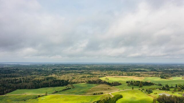 Aerial drone hyperlapse drone shot of white clouds drifting over rolling green fields, countryside forest, and a winding rural road.