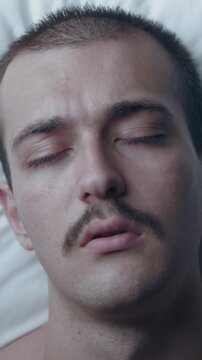 Top-down view portrait of a young Caucasian man with a mustache tossing and turning restlessly in bed, experiencing insomnia and bad nightmares on a white pillow.