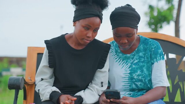 Black mother guiding daughter over smartphone, pointing at screen and explaining app, park bench setting, engaged eye contact, supportive teaching moment, informal tutoring between parent and child
