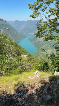 An upward camera tilt reveals Perucac Lake from Banjska Stena viewpoint on Tara Mountain, with rocky ground, dense forest, steep canyon slopes and a clear blue sky.