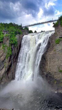 Vertical low angle view of Montmorency Falls surging down steep cliffs under suspension bridge in Quebec City, Quebec, Canada. 
