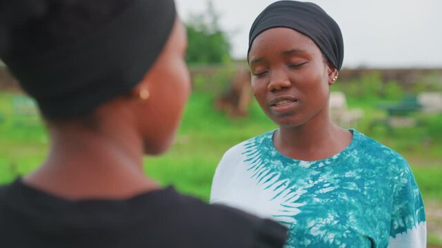 Black mother and daughter facing each other in rural garden, quiet confrontation, older woman wearing headwrap, younger figure turned away, subdued expressions, natural light, intimate portrait,
