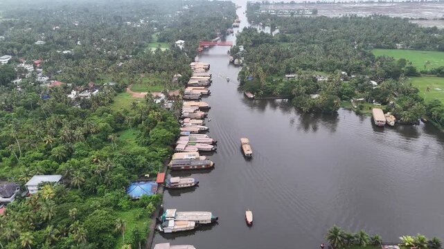 Journey towards Pathiramanal Island, a sanctuary in Vembanad Lake. Dozens of traditional houseboats dock along lush palm-fringed channels beneath a scenic red bridge in Alappuzha.