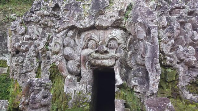 Pan right camera tracing the ornate carved demon face at the entrance of Goa Gajah, capturing centuries old Balinese stone craftsmanship overtaken by moss