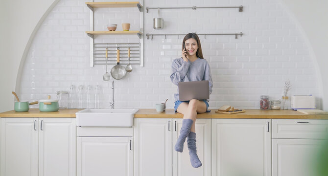 Portrait beautiful asian creative woman relax sit on kitchen counter use computer laptop phone meeting Happy asian girl at kitchen counter home. Freelance small business asian owner sme work from home