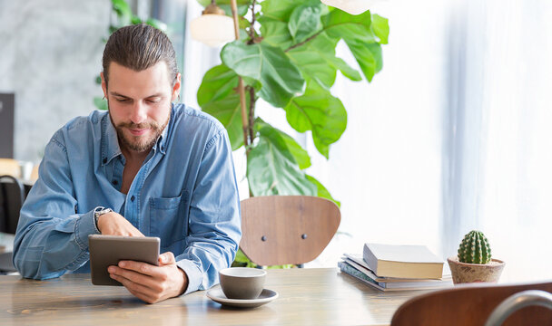 Young tattooed man sitting at a modern cafe, using a tablet near a window with coffee and a small cactus nearby. Relax slow life with technology