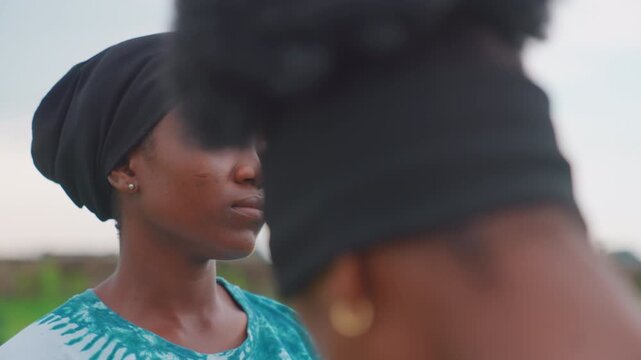 Black mother and child closeup outdoors, headscarf, eyes closed, reflective expression, blurred daughter foreground, green field background, intimate calm with undercurrent of tension and compassion,