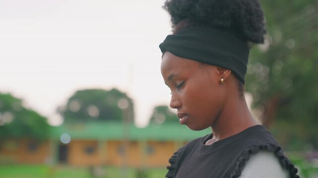 Young woman in profile outdoors, looking down in reflective mood with headwrap and textured sweater. Soft evening light, blurred green background, calm composition, subtle emotion, candid student