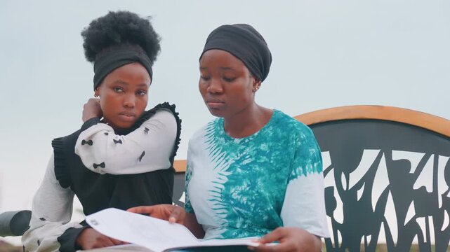 Black mother and child studying outdoors, on patterned bench, reviewing papers, mother pointing and explaining, daughter listening with concentration, intimate tutoring moment, homework focus, calm