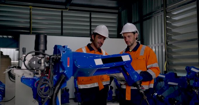 Two adult male technicians monitor industrial robotic arm movement, reviewing handheld controller and discussing equipment operation during collaborative automation work inside factory workshop.