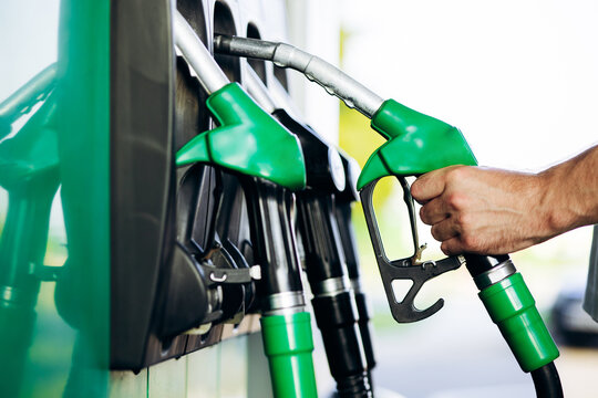 Person's hand pulling the trigger on a green fuel pump nozzle at a gas station, filling a vehicle with gasoline