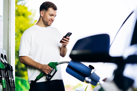 Man refilling vehicle with gasoline, holding a fuel pump nozzle and checking his mobile phone at a gas station