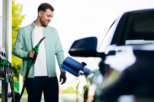 Man holding a green fuel nozzle, filling his car with gasoline at a modern service station, looking down with a smile