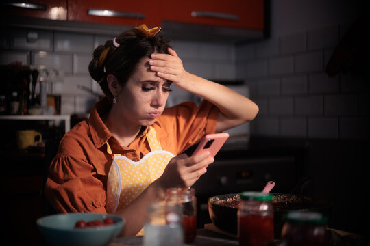 Exasperated Housewife Looking At Phone Recipe While Cooking Homemade Jam. Stressed young woman checking smartphone recipe in kitchen holding hand on forehead