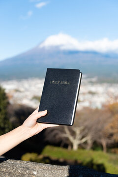A hand holds a black, hardcover Holy Bible aloft with the iconic snow-capped Mount Fuji and Fujinomiya city in the background, from the Shirao Mountain Park (Shiraosan), Japan. Verical shot.