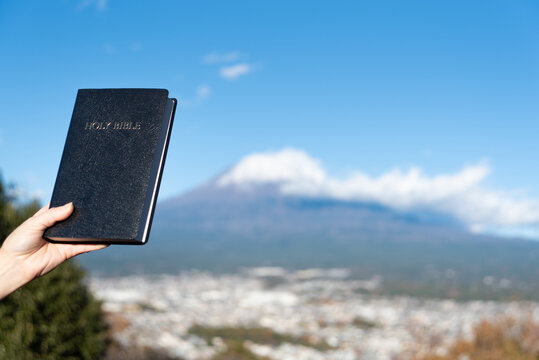 A hand holds a black, hardcover Holy Bible aloft with the iconic snow-capped Mount Fuji and Fujinomiya city in the background, from the Shirao Mountain Park (Shiraosan), Japan. Horizontal shot.