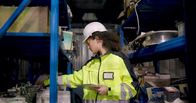Adult female technician uses tablet while checking spare parts on storage rack in factory workshop, comparing equipment components during automation maintenance planning and inventory inspection.
