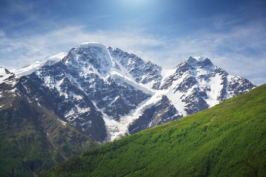 Landscape of a high rocky mountain with massive snowy peaks and glacier slopes under a blue sky with soft white clouds.