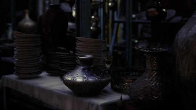 Dimly lit market table with pottery, closeup stacked plates and vases in shadowy medina stall, textured glaze and earthen tones, lantern highlights on patina