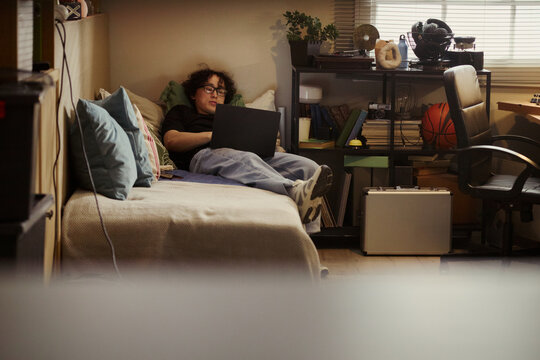 Asian young man lying on bed and using laptop in messy bedroom, studying or gaming. Image represents youth digital lifestyle, suitable for remote learning and student housing campaigns