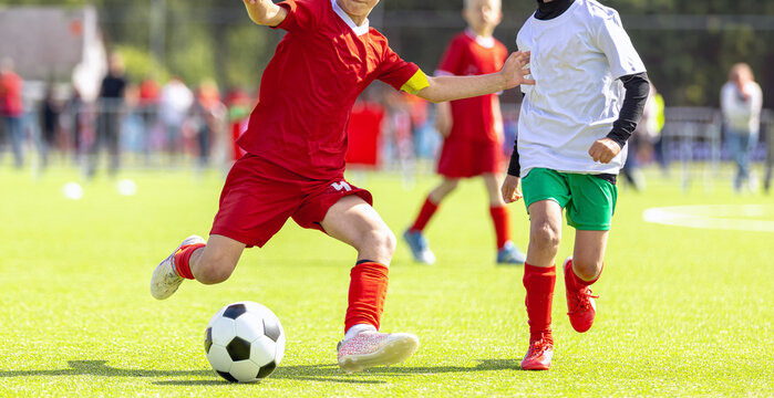Junior Soccer Player in Red Kit Kicking Ball Past Defender - Competitive Youth Football League Match Action on Sunny Grass Pitch