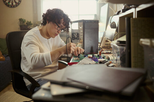 Asian young man soldering motherboard at desk, repairing computer hardware. Image represents engineering education, IT support services, and DIY technology repair, medium shot