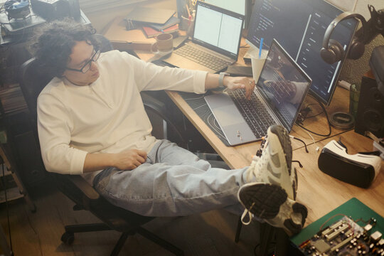 Young Asian man coding on laptop with feet on desk, working in home office. High angle view of software developer programming, suitable for remote work and IT career concepts
