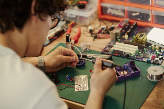 Asian young man soldering circuit board at workshop desk, learning electronics engineering. Image suitable for STEM education campaigns and youth technology training programs