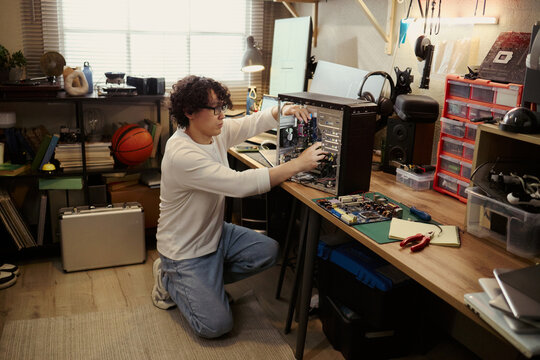 Young Asian man repairing desktop computer hardware in home workshop. Image represents IT support, technology maintenance, and DIY electronics repair for tech blog use