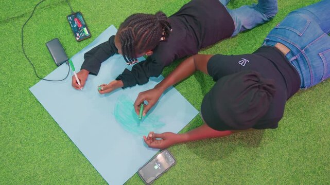 Black mother and child creating outdoors on grass with blue sheet, collaborative doodling and coloring, mother as creative partner, playful gestures, phone and charger visible, casual jeans, braided