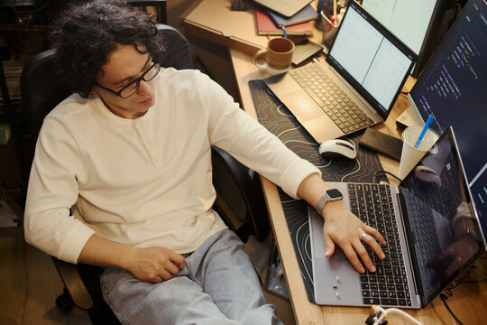 Young Asian man writing code on laptop at desk with multiple screens, representing software development and remote work. High angle shot