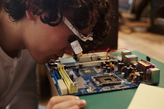 Asian young man wearing magnifying glasses examining computer motherboard. Image represents STEM education and hardware engineering, suitable for technology training school marketing