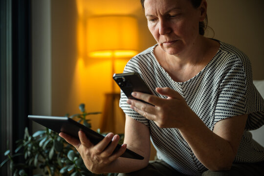 Mature woman using a digital tablet and smartphone to synchronize data between devices at home. Cross-device connectivity, cloud technology and digital productivity concept.