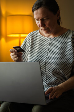 Middle-aged woman making an online purchase with a laptop and bank card in a cozy living room. Internet shopping, cashless payment and home convenience theme.