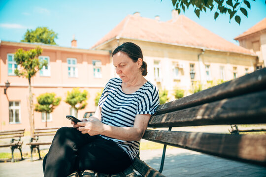 Mature woman relaxing on a park bench while browsing her smartphone beneath leafy trees. Urban lifestyle, connectivity and everyday leisure theme.