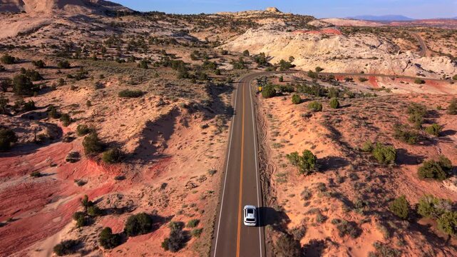 Aerial drone footage Utah Scenic Byway 12 shows white car traveling along winding desert highway. Red sandstone formations and scattered juniper trees create stunning landscape under clear blue sky.
